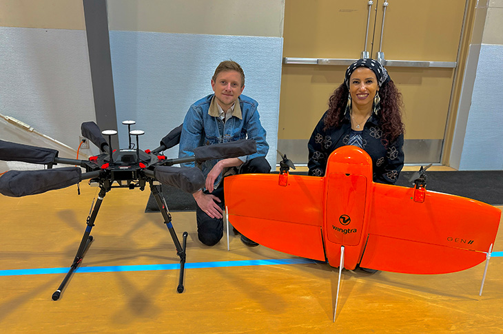 Two people crouch behind two drones in a gym, one is a multi-rotor black device and the second is an orange fixed wing.