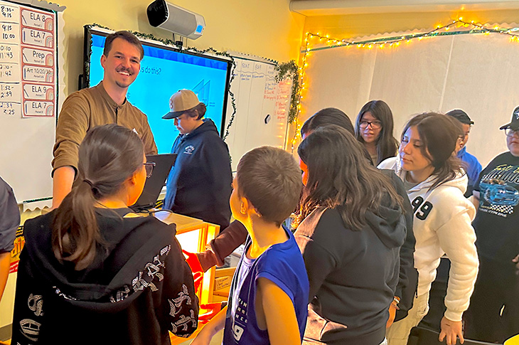 A man stands at the front of a classroom smiling at the camera and surrounded by students.