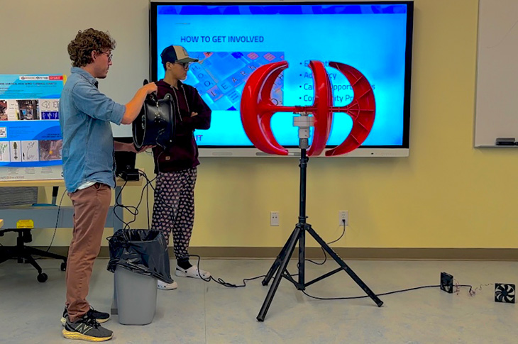 Two people stand at the front of a classroom in front of a screen. One points a fan at a small horizontal-axis wind turbine.