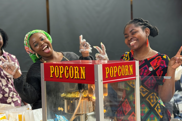 Two students smile and make the peace sign with their hands while standing behind a popcorn machine at Black History Month Fest 2025.