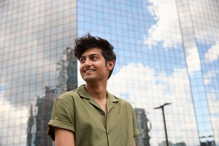 smiling SAIT student looks into the distance with tall glass-clad buildings in the background reflecting other buildings, blue sky, grey and white clouds