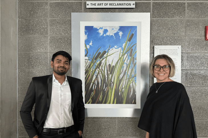 A SAIT student in a suit and an adult woman in a long-sleeved black shirt stand in front of a painting. The painting features tall cattails swaying in the windy against a bright blue sky. Above the painting, a sign reads "The Art of Reclamation."