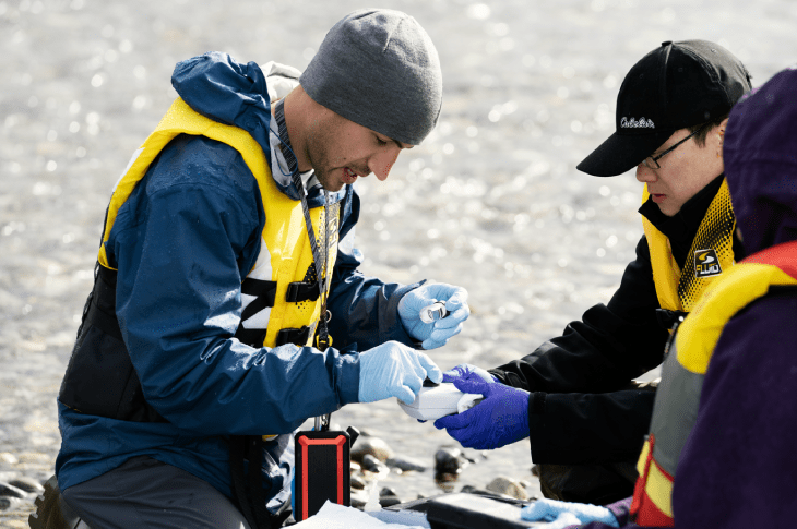Two students in safety vests and gloves conduct water quality testing by a riverbank.