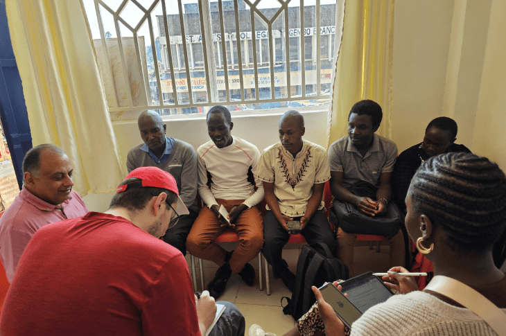 A group of six people seated together in a well-lit room, engaged in conversation, while another person takes notes.