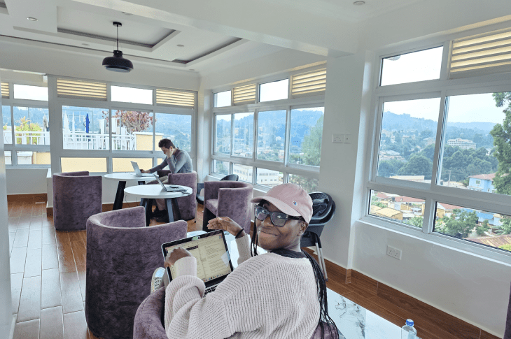 A student in a pink cap sits at a table working on her laptop in a modern, bright workspace, with a view of mountains in the background.