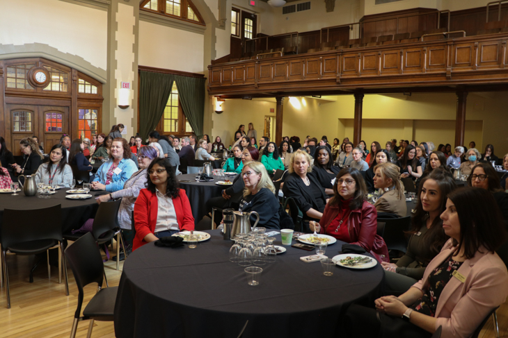 A diverse group of attendees sitting at round tables during an event, engaged and listening attentively.