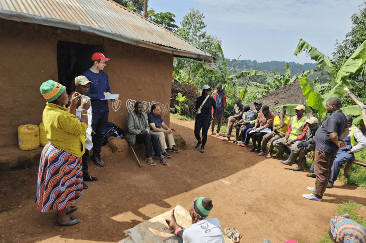 A community meeting taking place outside a mud-brick house, with attendees sitting and standing among tropical plants.