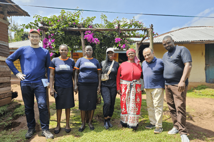 A group of six individuals, including community members and visitors, standing together in front of colorful flowers and greenery in a rural setting.