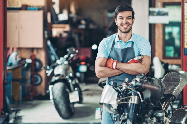 A young man wearing safety gloves and apron crosses his arms and smiles at the camera, while standing in a motorcycle repair shop.