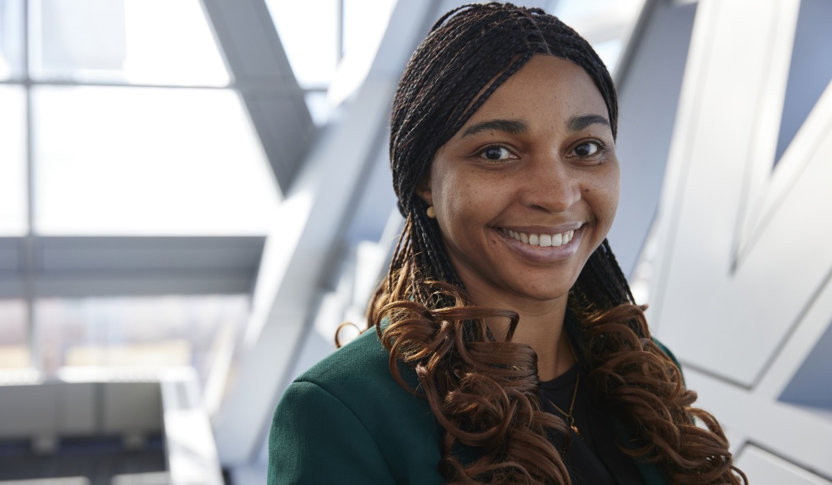 A smiling woman with braided hair, wearing a green jacket, stands in a modern architectural space with large windows.