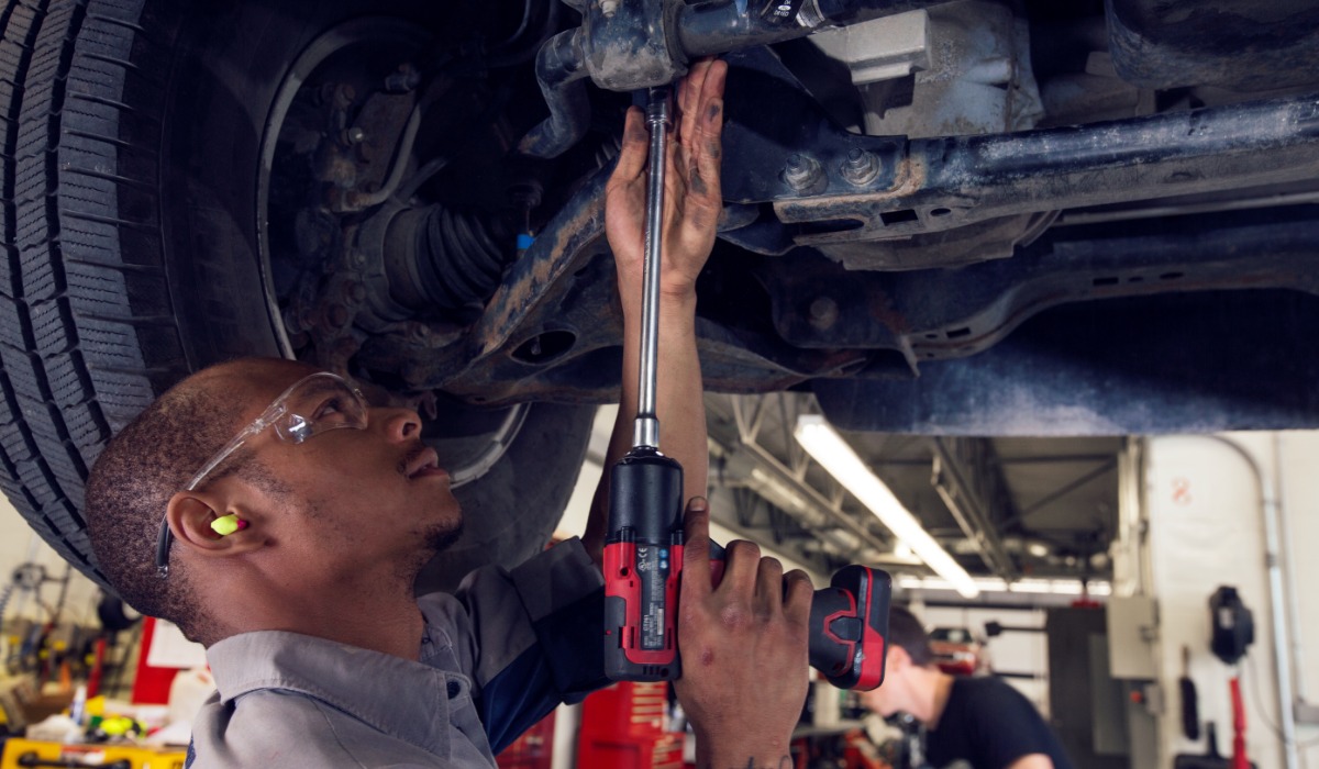 A mechanic using a power tool to inspect or repair the undercarriage of a vehicle in a workshop.