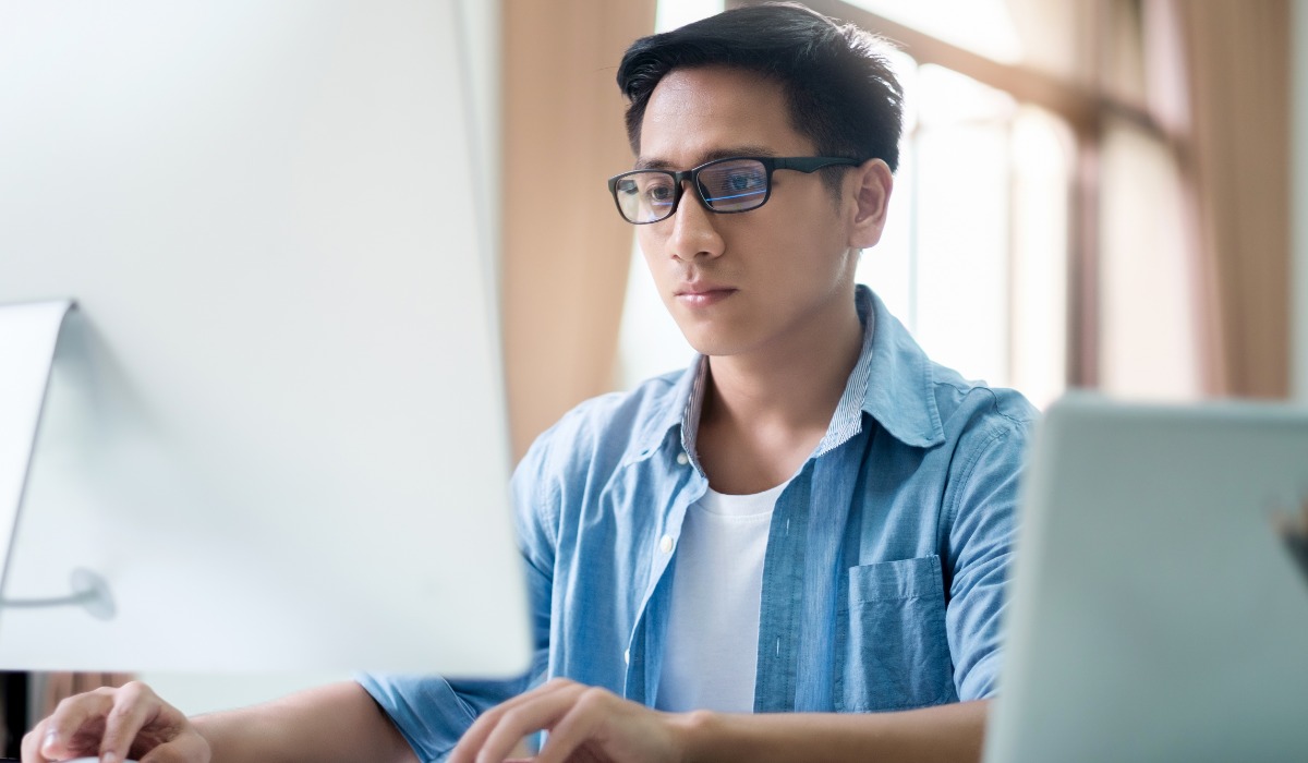 A young man wearing glasses focuses intently on a computer screen while working at a desk.