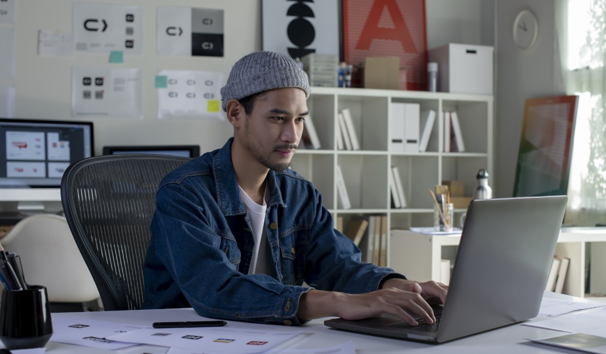 A young man in a denim jacket and beanie working on a laptop in a modern office space filled with design elements and sketches.