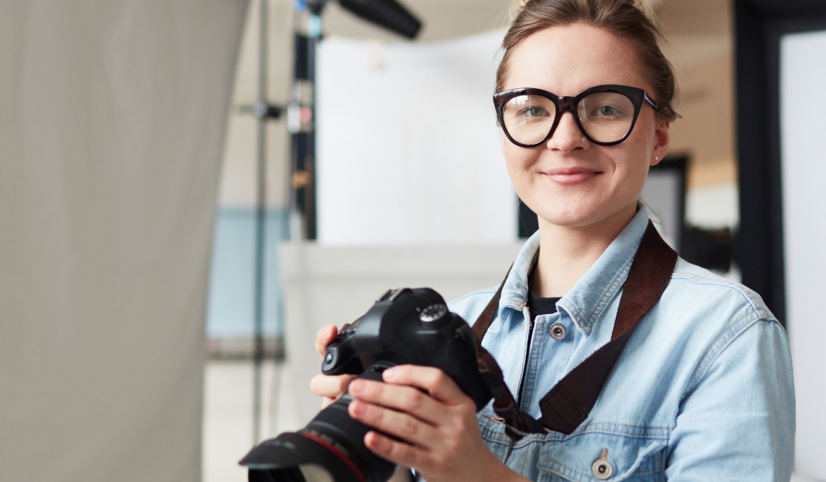 A smiling female photographer wearing glasses and a denim jacket, holding a camera.
