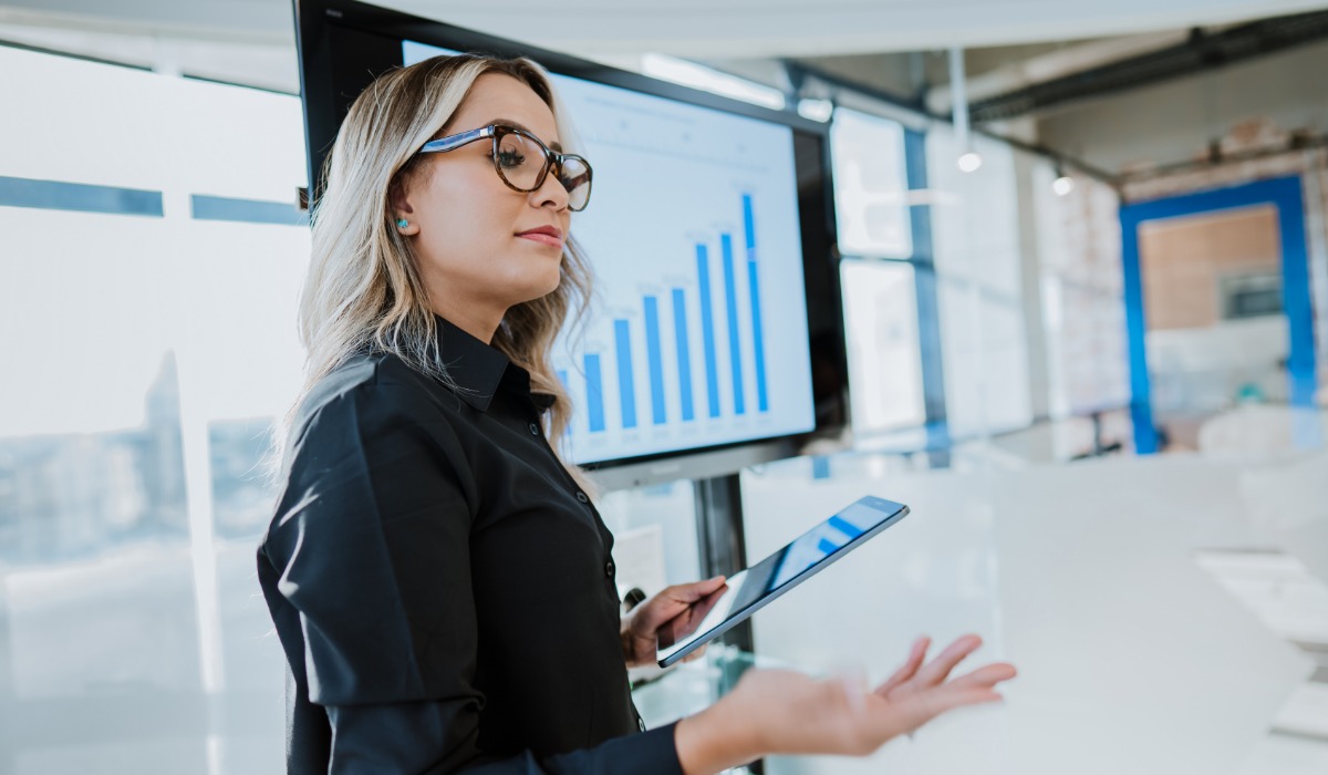 Businesswoman presenting data with a tablet in front of a screen displaying bar graphs.