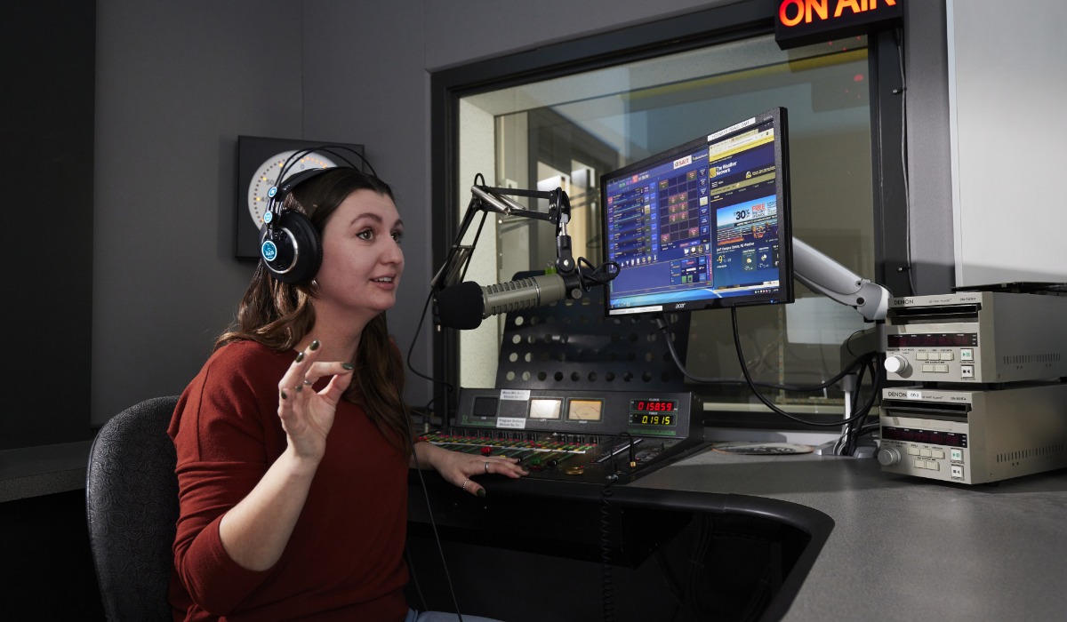 Radio host speaking into a microphone while operating a soundboard in a broadcast studio.