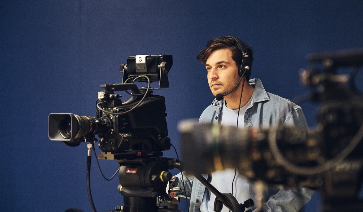 A young male videographer adjusts a professional camera while wearing headphones in a studio setting.