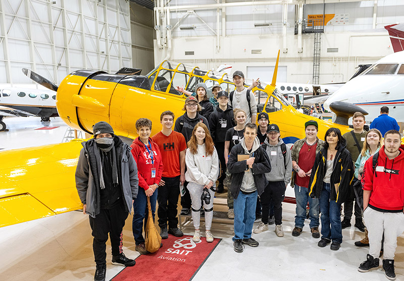 A group of young high school students standing and smiling in front of a small yellow aircraft 